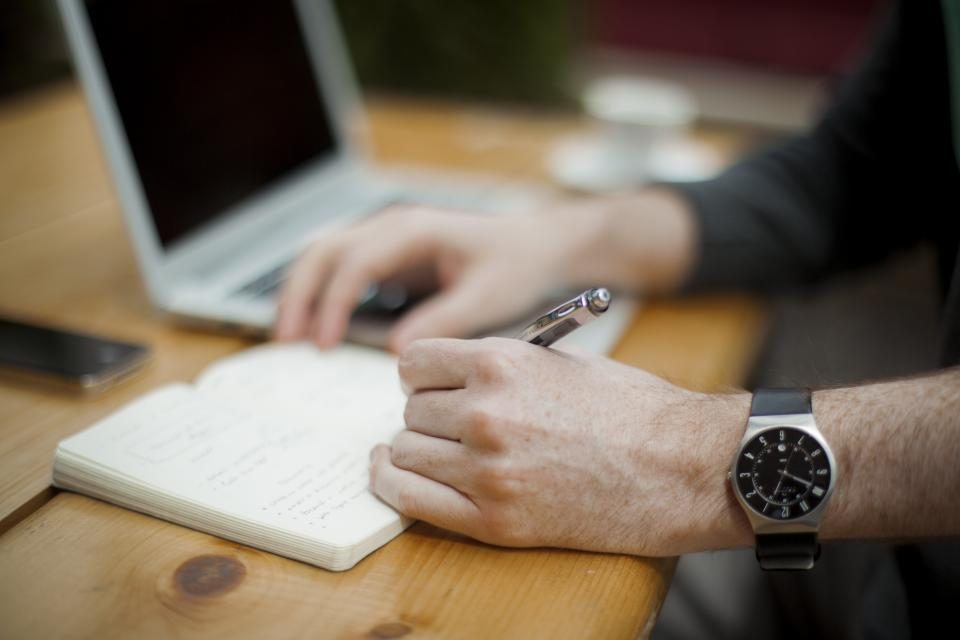 Hands writing in a notebook beside a laptop