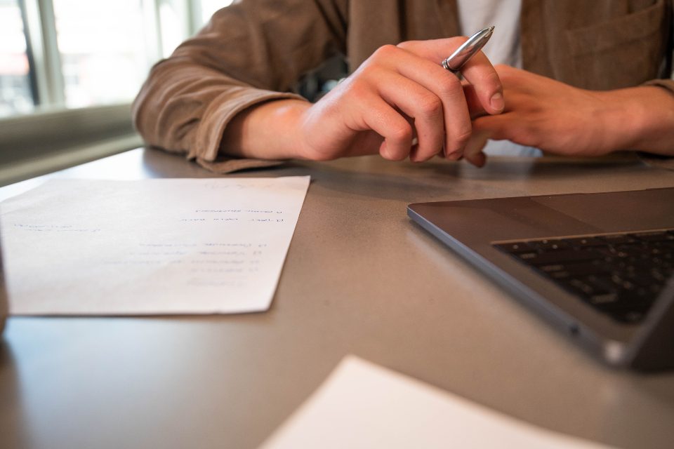 Person with a pen reviewing papers beside a laptop