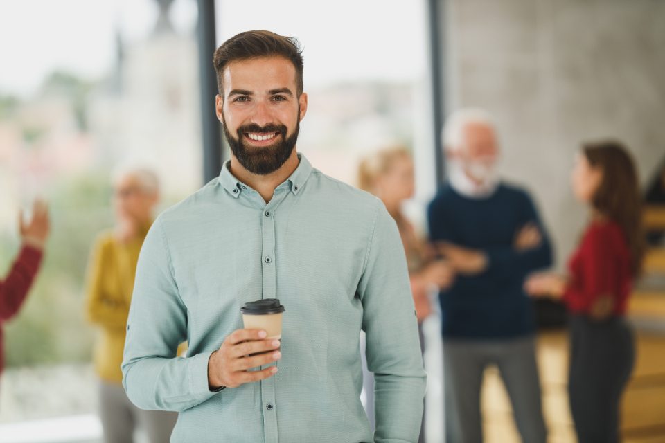 Smiling man holding a coffee with colleagues behind him